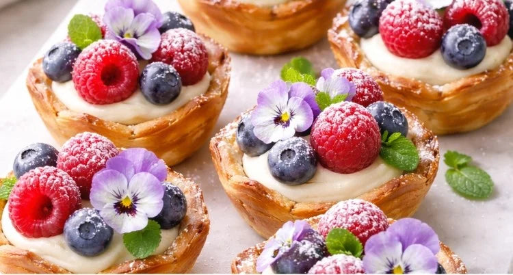 Six mothers day tart shells filled with whipped vanilla cream and topped with fresh raspberries, blueberries, and purple pansies, styled on a white marble board.