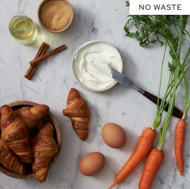 Ingredients for a no-waste carrot cake, including croissants, fresh carrots, eggs, cream cheese, cinnamon sticks, brown sugar, and oil on a marble countertop.